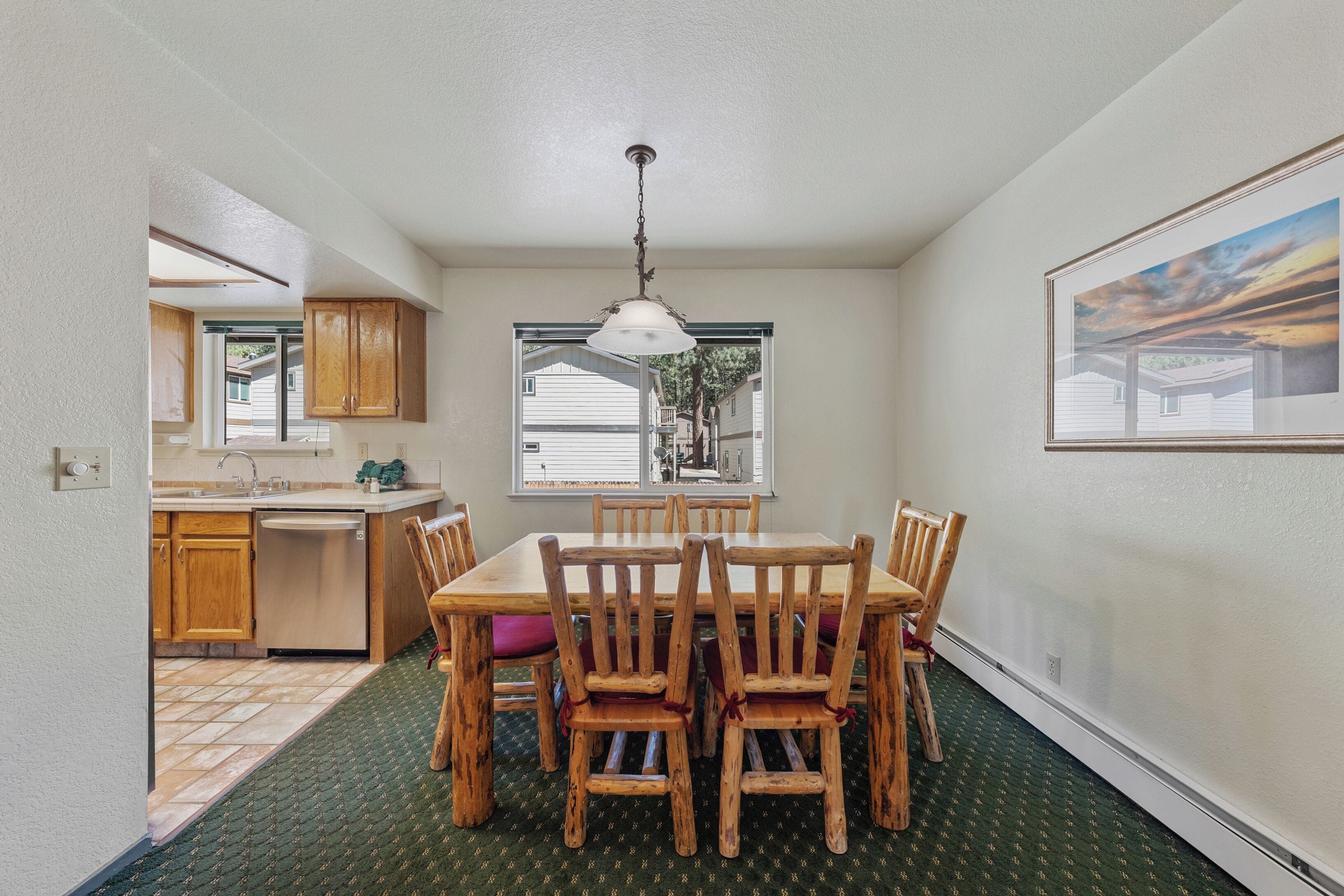 A dining area with a wooden table and chairs, a window with a view of houses, and a kitchen with wooden cabinets.