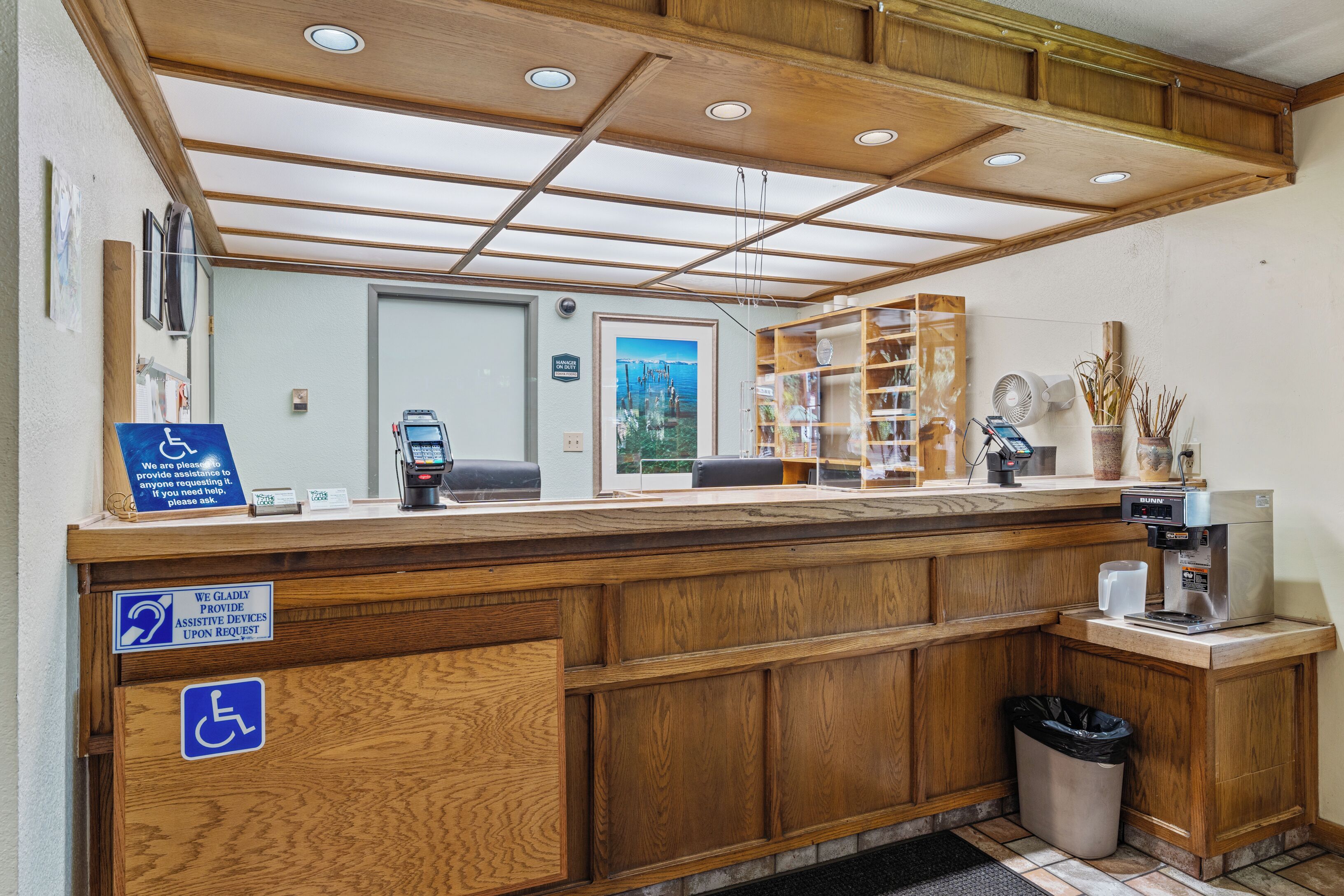 A reception area with a wooden counter, a trash bin, a coffee machine, and a sign indicating the availability of assistive devices upon request.