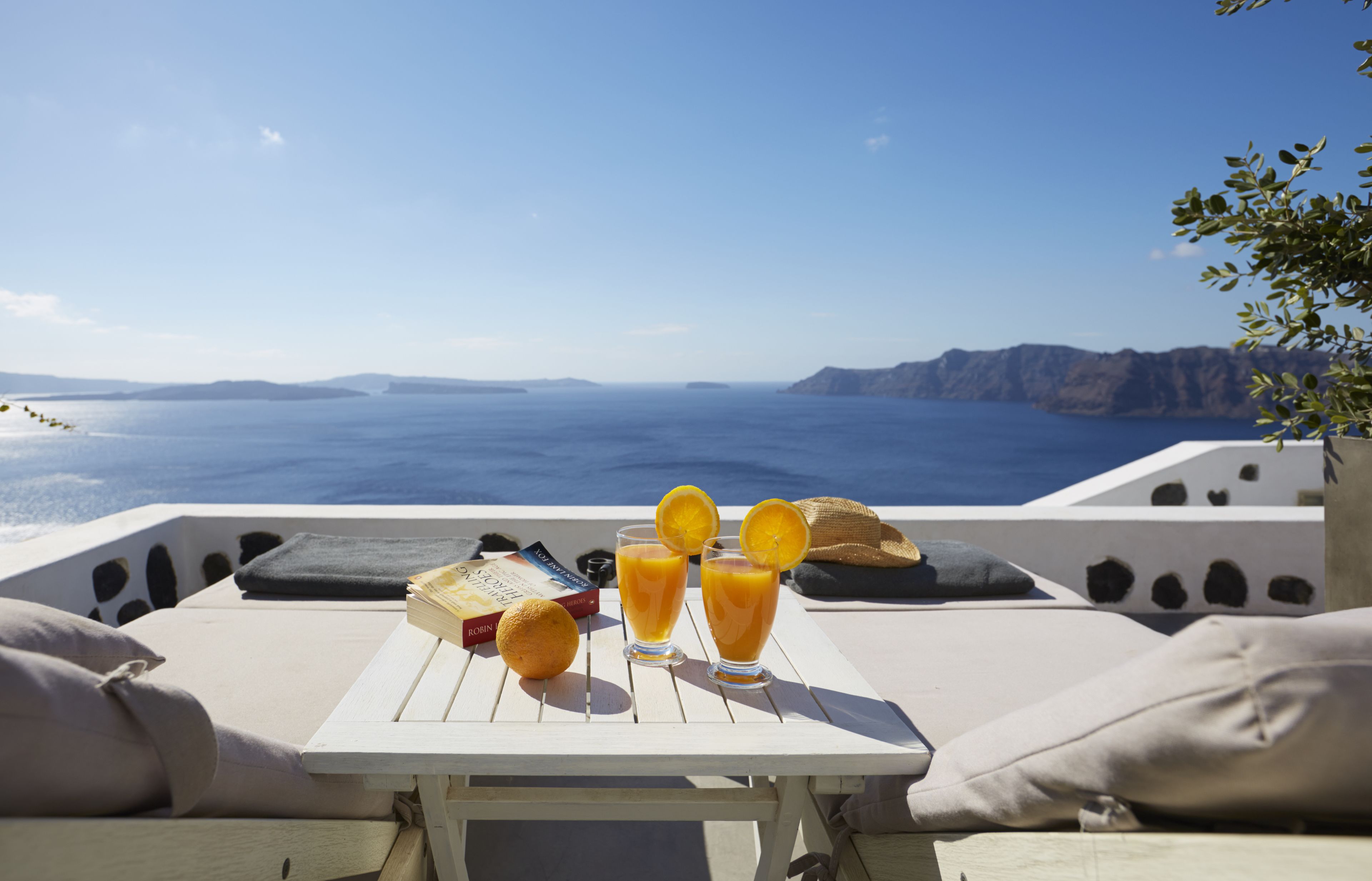 Une table avec un livre, deux verres de jus d’orange et un fruit, sur une terrasse offrant une vue sur la mer et les montagnes lointaines.