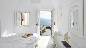 A bright, white room with a bed, a table with a sculpture, and a view of the sea through an open door. - Atrina Canava 1894 (Santorini)