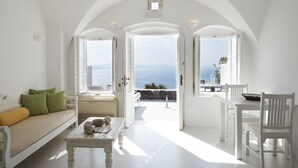 A bright, white interior with a sofa, table, and dining area leading to a balcony with a view of the sea. - Atrina Canava 1894 (Santorini)
