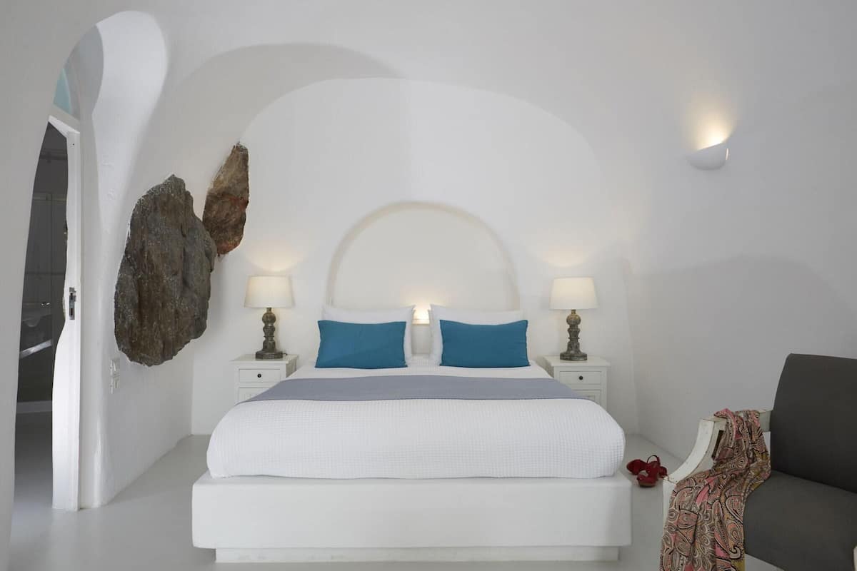 Quarto com cama branca, duas almofadas azuis, mesa de cabeceira branca com abajur, cadeira cinza e uma grande decoração de rocha na parede.