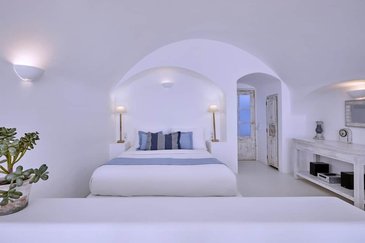 Quarto minimalista com uma cama grande, paredes brancas e uma porta em arco que dá para outro cômodo.