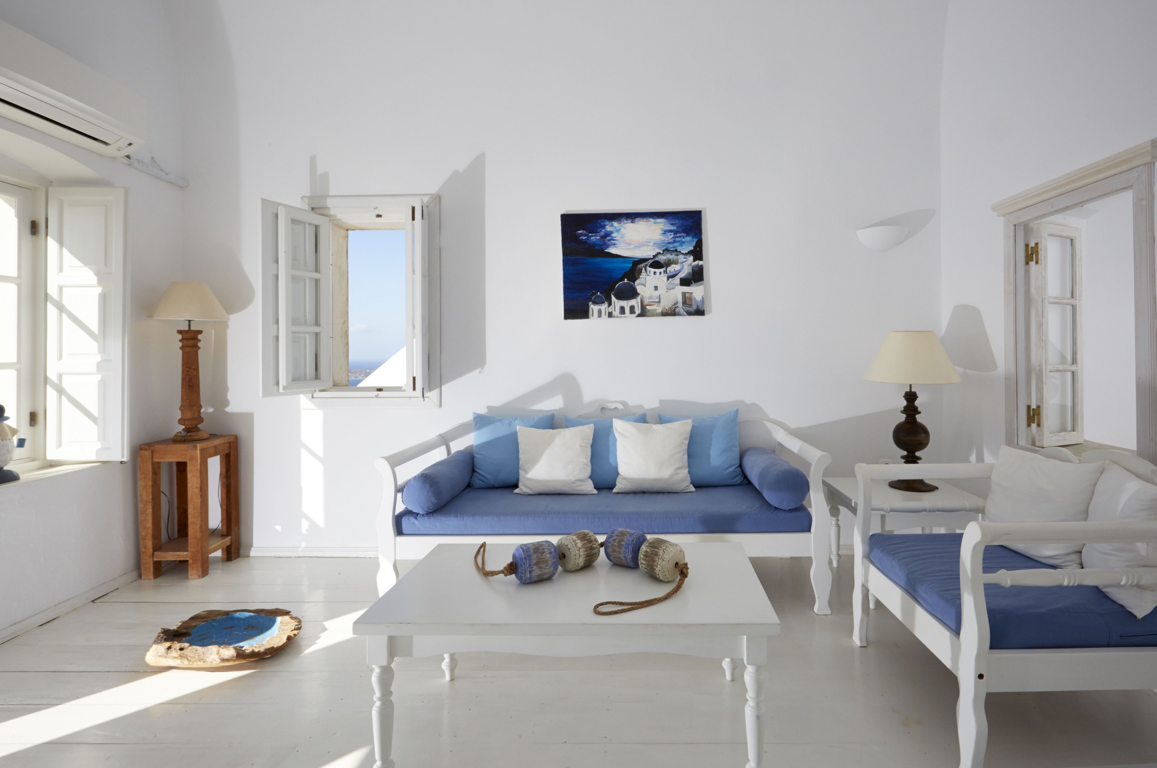 A room with a white sofa, a small table, and a bed with blue bedding. There is a painting on the wall and a lamp on the table.