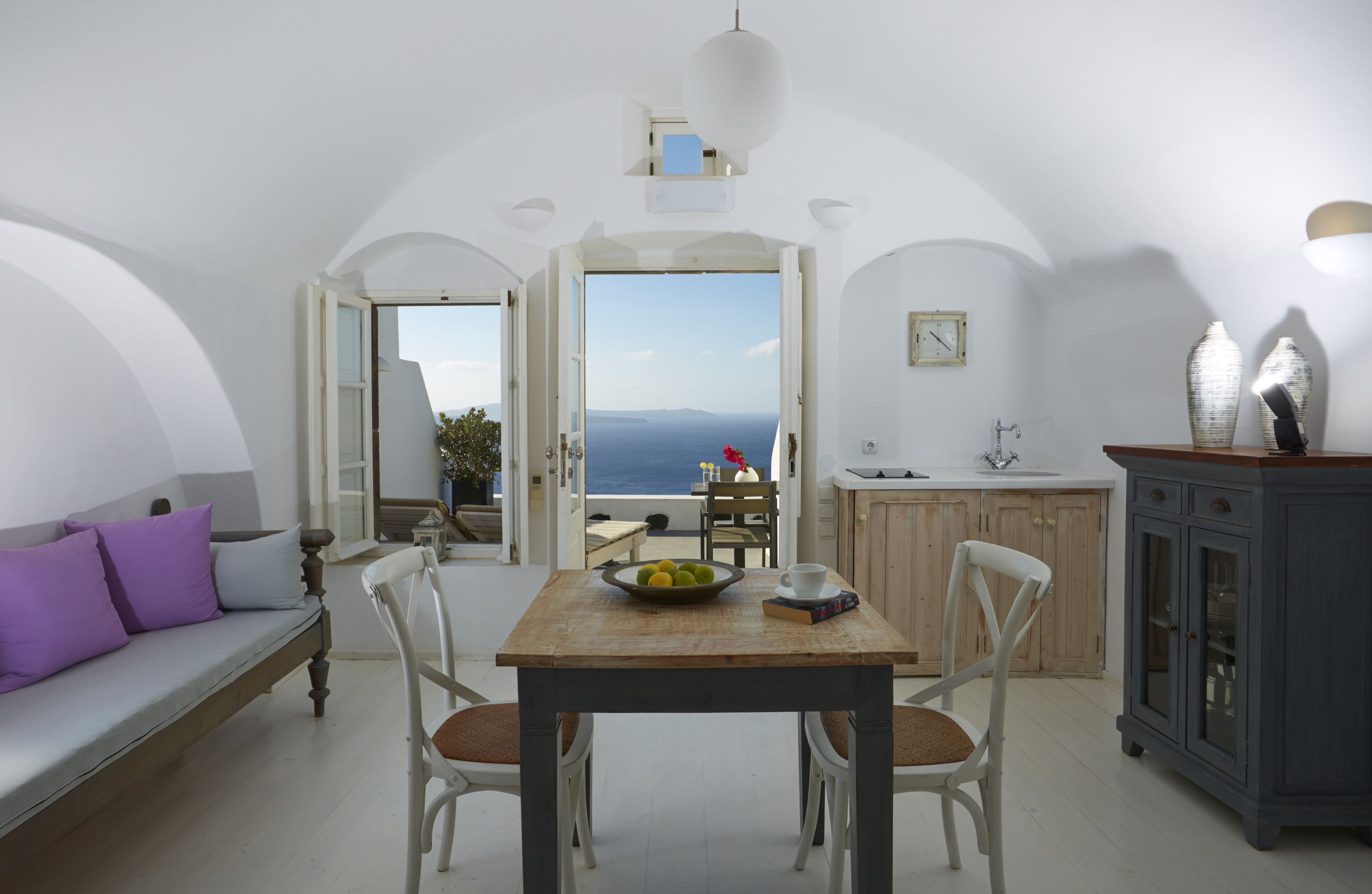 A room with a wooden table, white chairs, a sofa with purple cushions, a kitchen area, and a view of the sea.