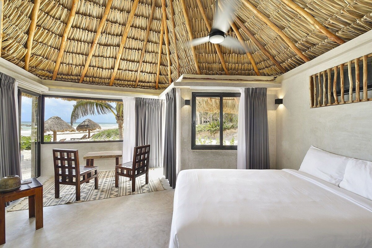 A room with a bed, a table, chairs, a ceiling fan, and large windows with curtains.