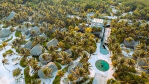 Vista aérea de un complejo turístico con cabañas de techo de paja, palmeras y una piscina.