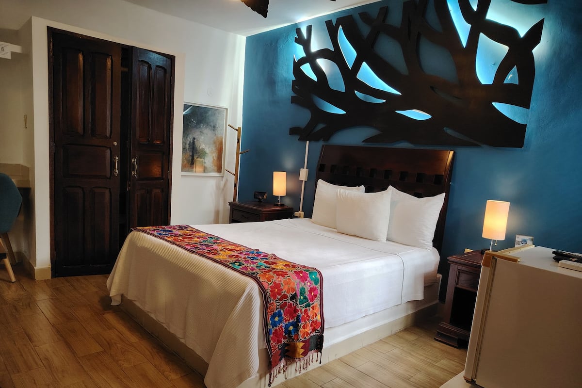 A hotel room with a bed, wooden floor, a blue accent wall, and a large tree decoration.