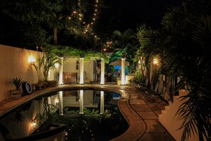 Zona de piscina nocturna con piscina circular, palmeras e iluminación ambiental.
