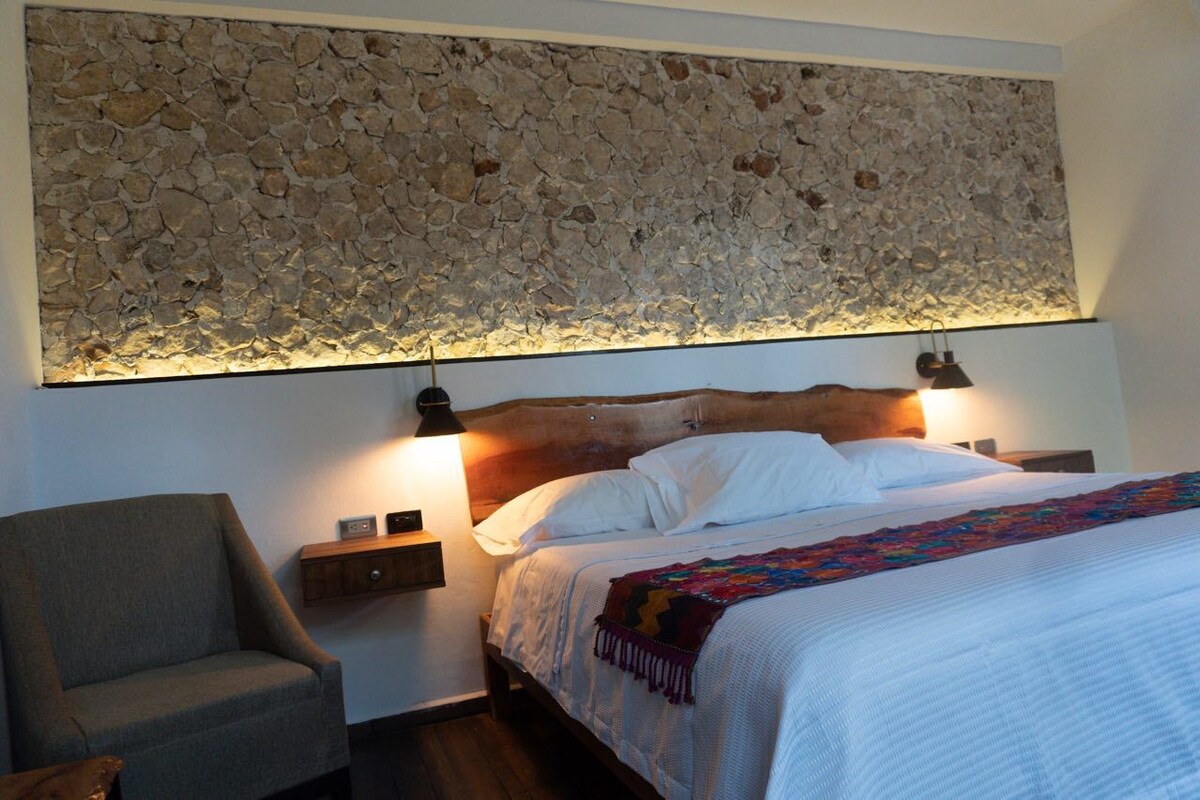 Un dormitorio con pared de piedra, cama con ropa blanca, dos lámparas de noche y una silla.