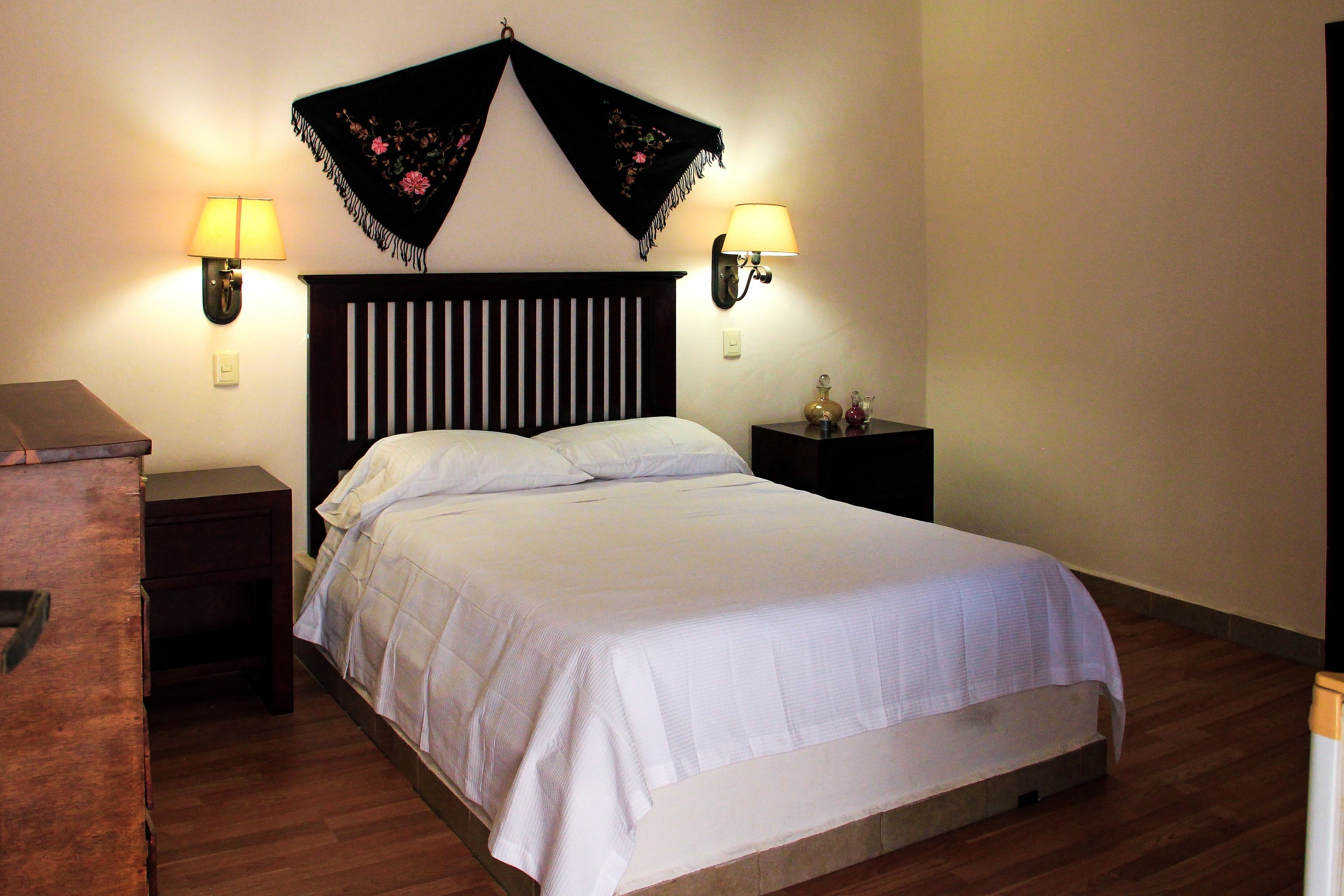 A neatly made bed with white linens, two bedside lamps, a dark wooden headboard, and a small nightstand with a decorative item.