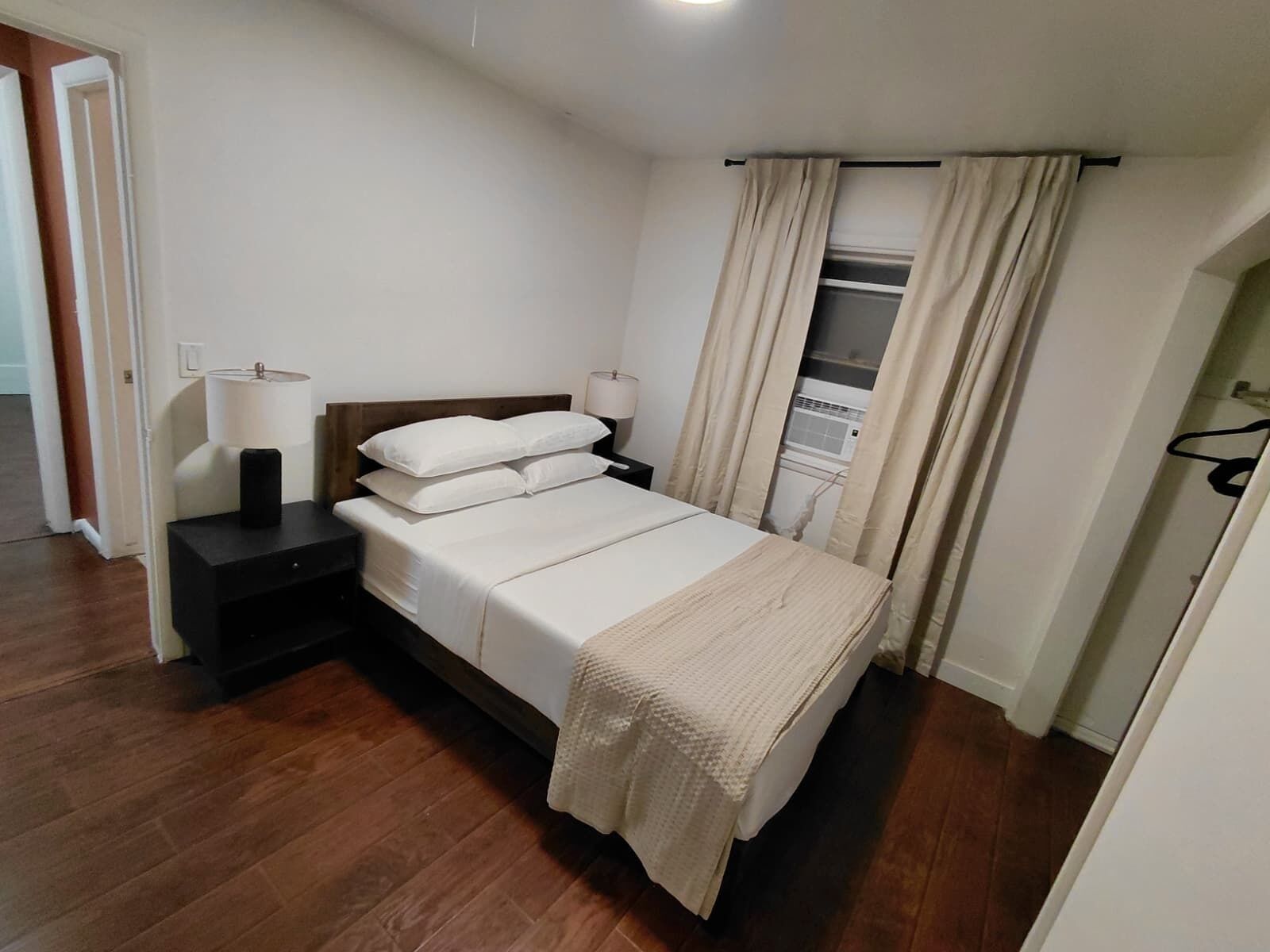 A bedroom with a bed, bedside lamps, a wooden headboard, a window with curtains, and a wardrobe.