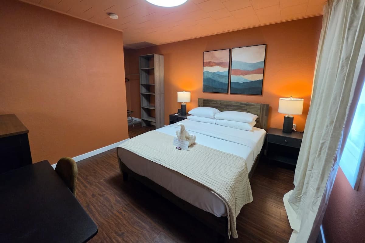 A hotel room with a bed, two bedside lamps, a nightstand, a desk, a chair, a wardrobe, and a painting on the wall.