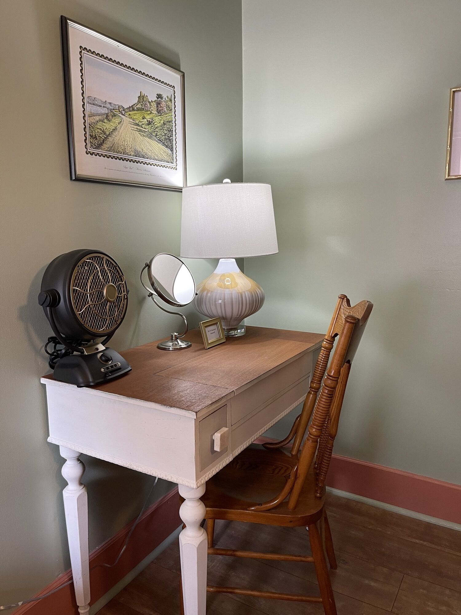 A desk with a lamp, a fan, and a mirror. There is a chair and a framed picture on the wall.