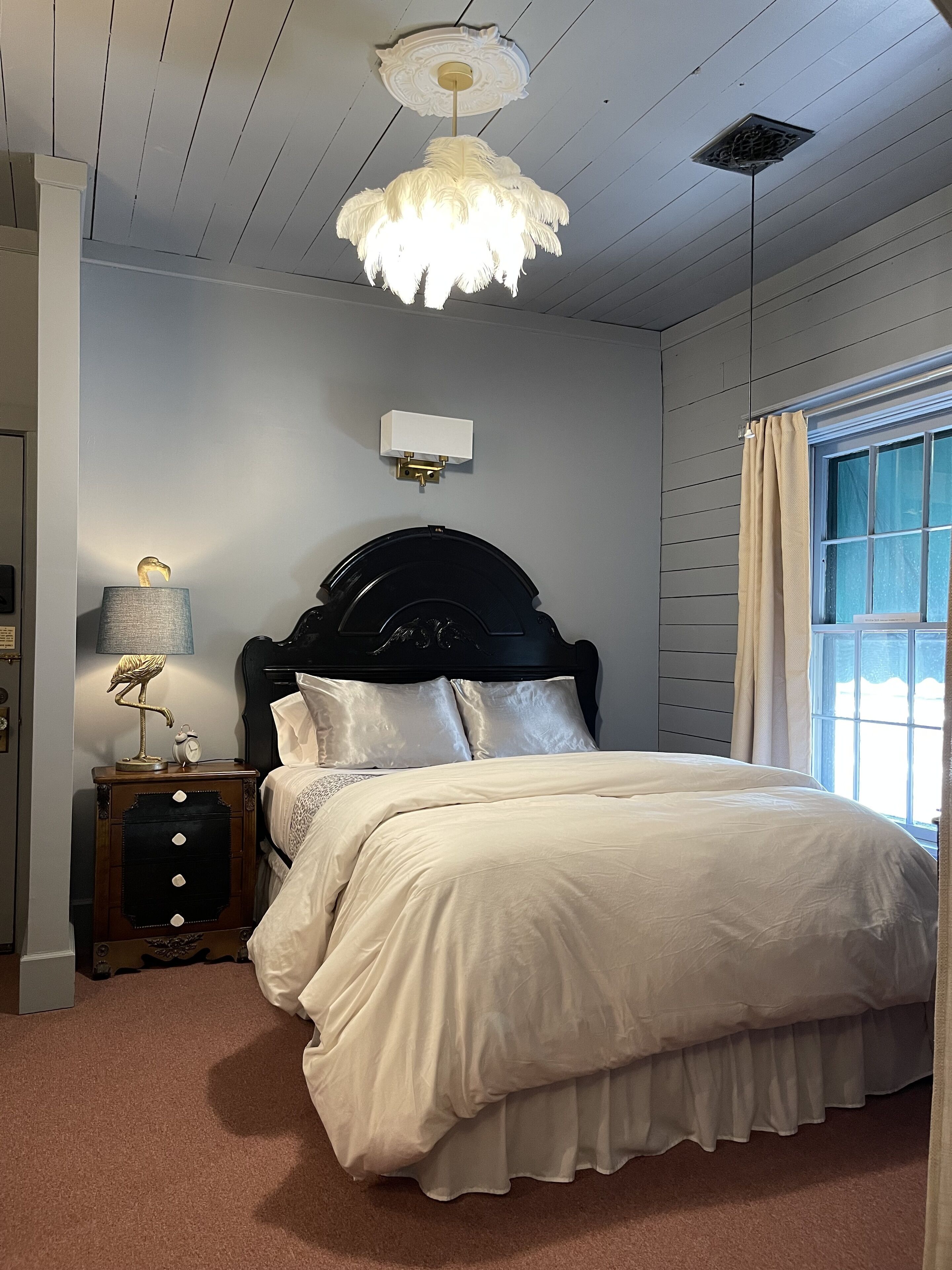 A bedroom with a bed, a nightstand, a lamp, a wall clock, and a window with curtains.