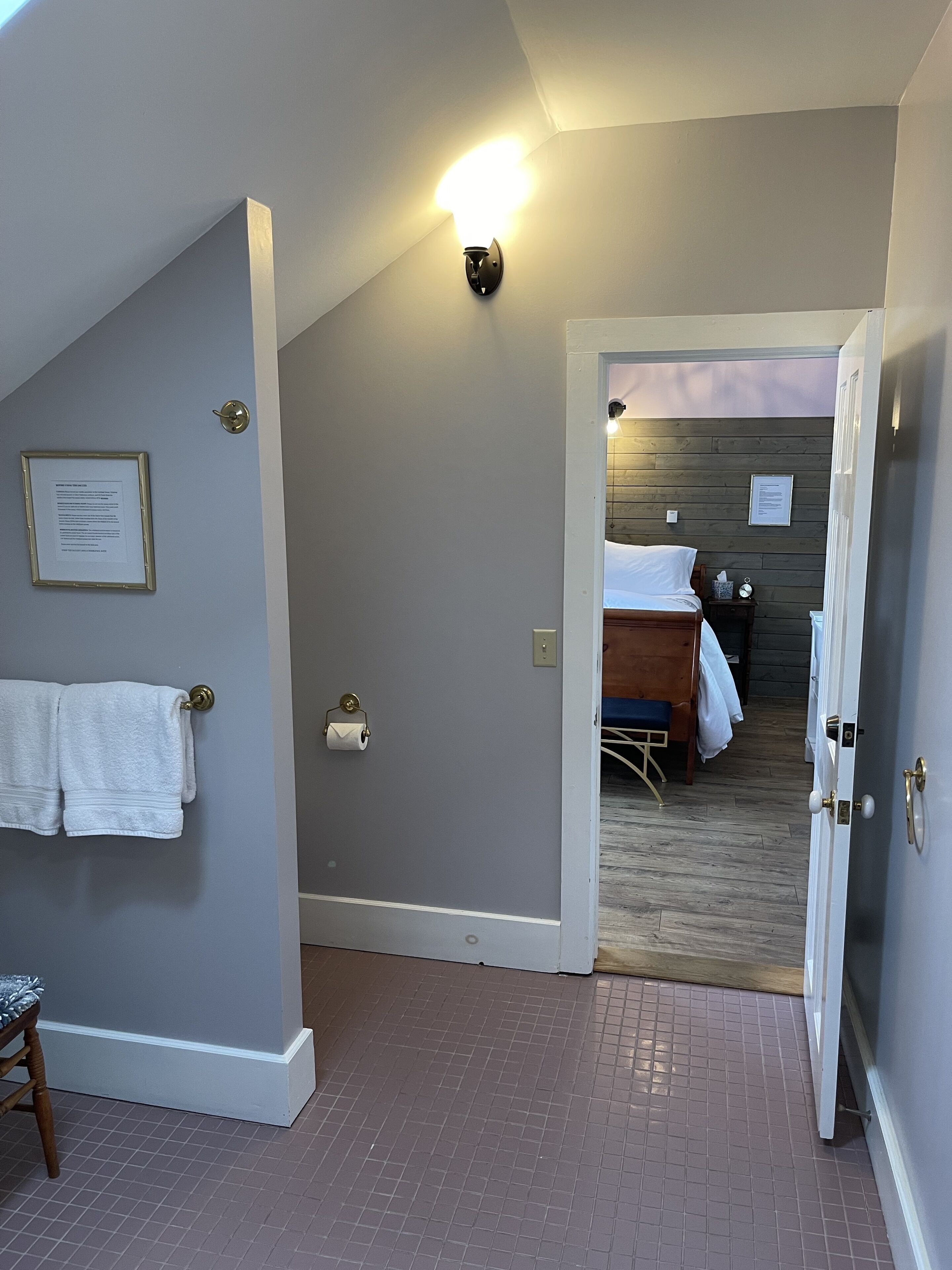Een hotelkamer met een bed, een klein tafeltje, een stoel, een handdoekrek en een wandlamp.