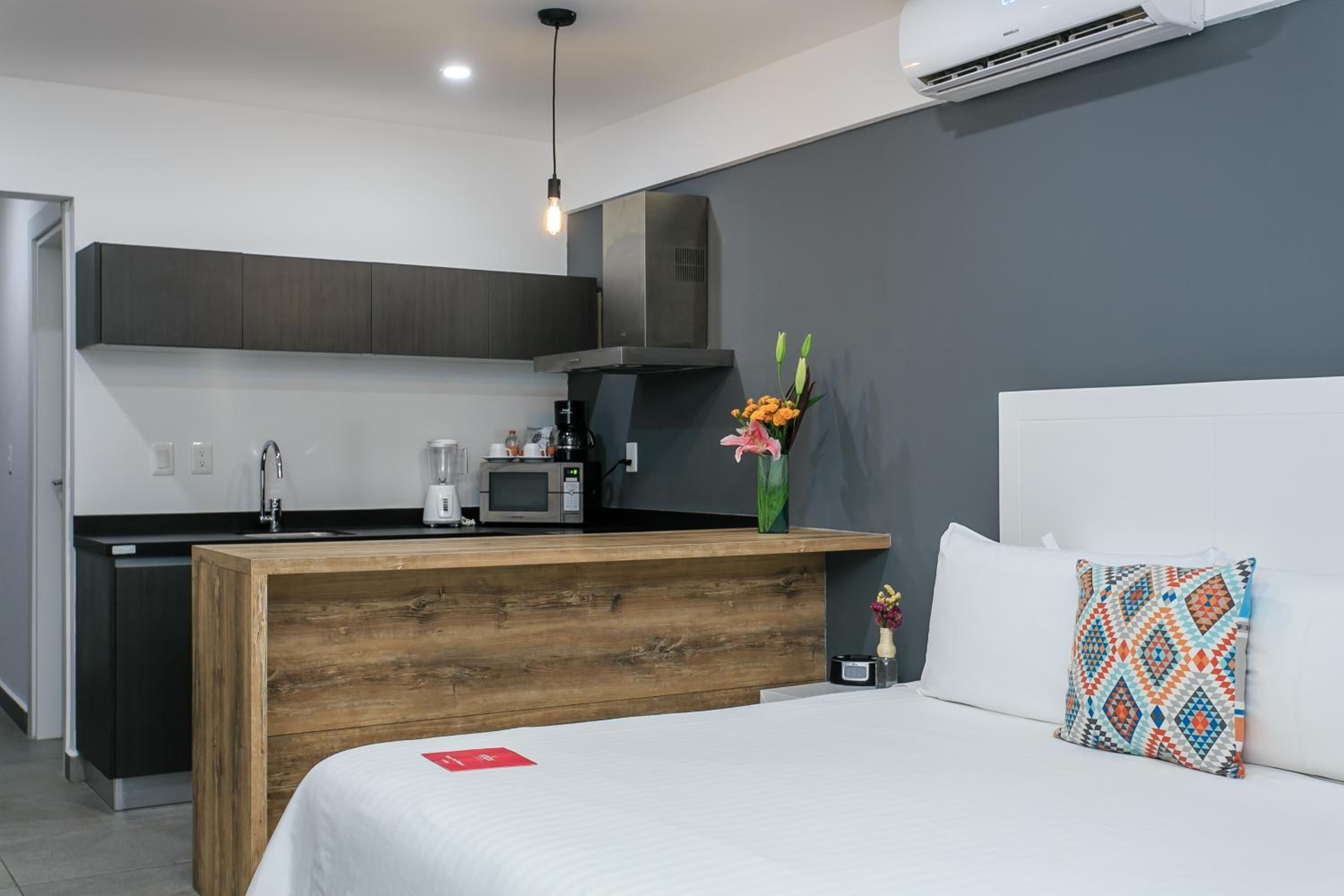 A modern hotel room with a wooden headboard, a white bed, a small kitchenette, and a grey accent wall.
