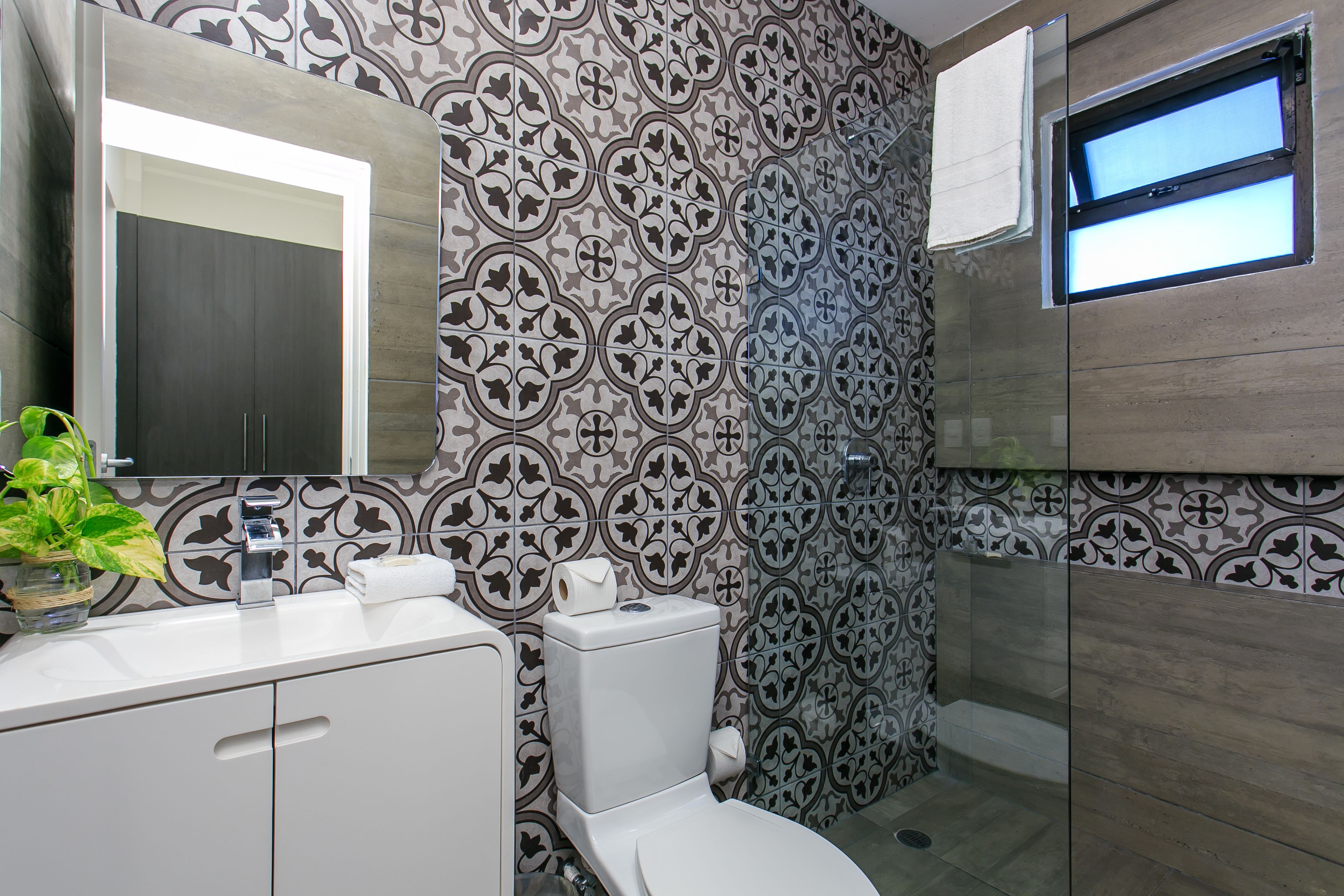A modern bathroom with patterned tiles, a toilet, a sink with a mirror, and a window.