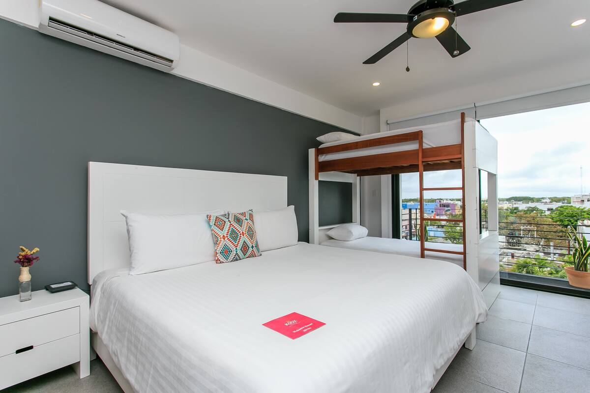 A modern hotel room with a large bed, a white nightstand, a ceiling fan, and a view of the city through large windows.