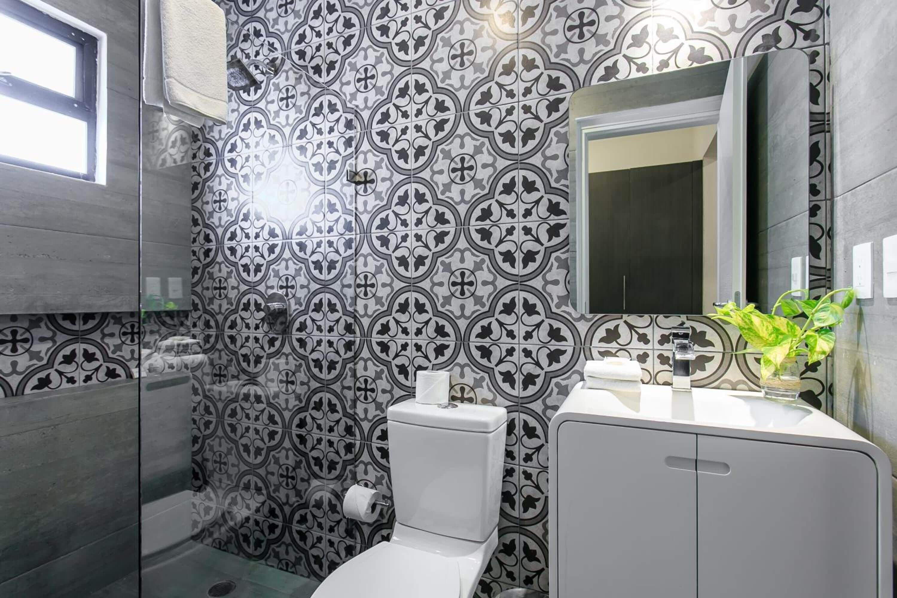 A bathroom with a patterned tile wall, a toilet, a sink with a mirror, and a window.