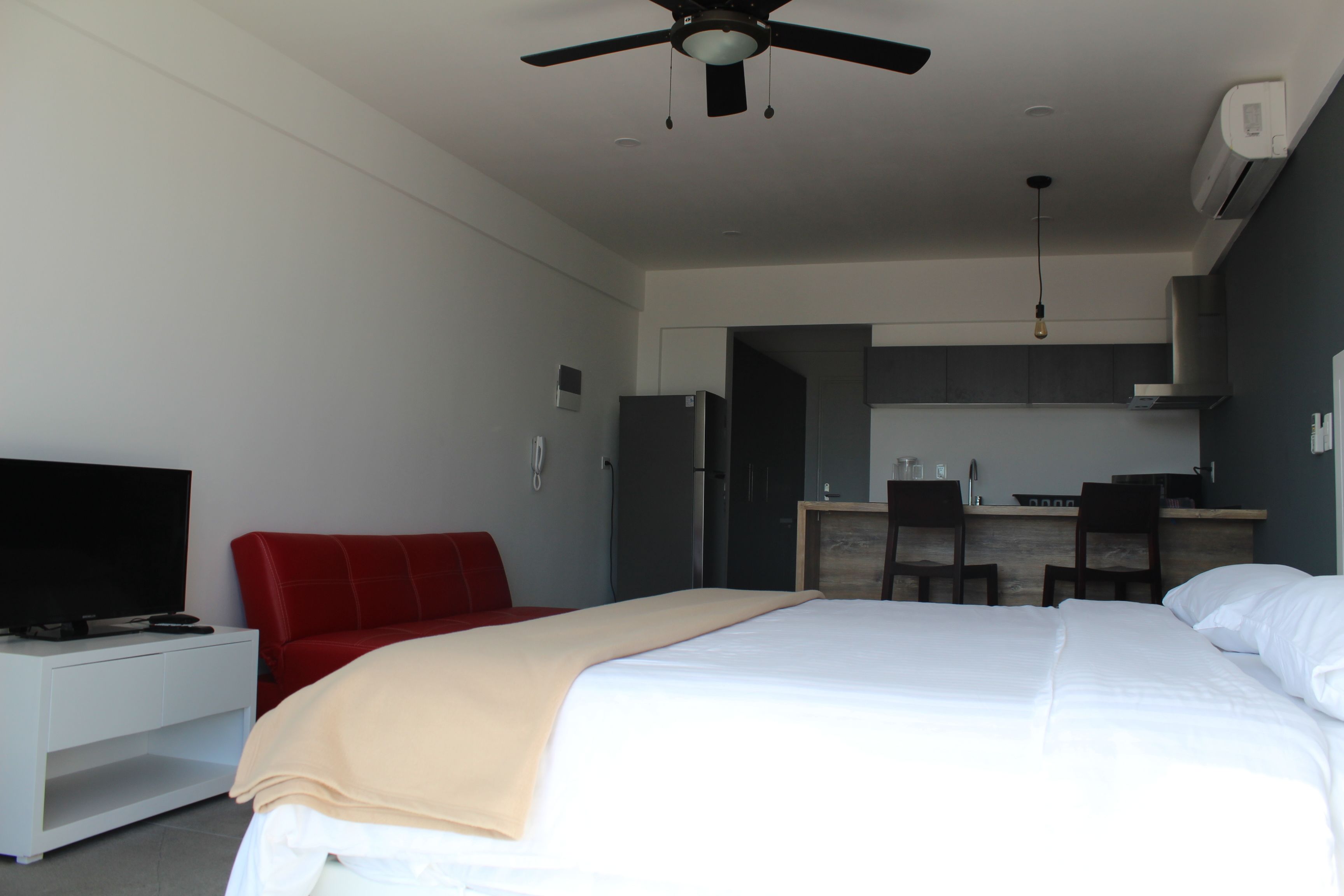 Una habitación de hotel moderna con cama, televisión, una silla roja y una kitchenette al fondo.