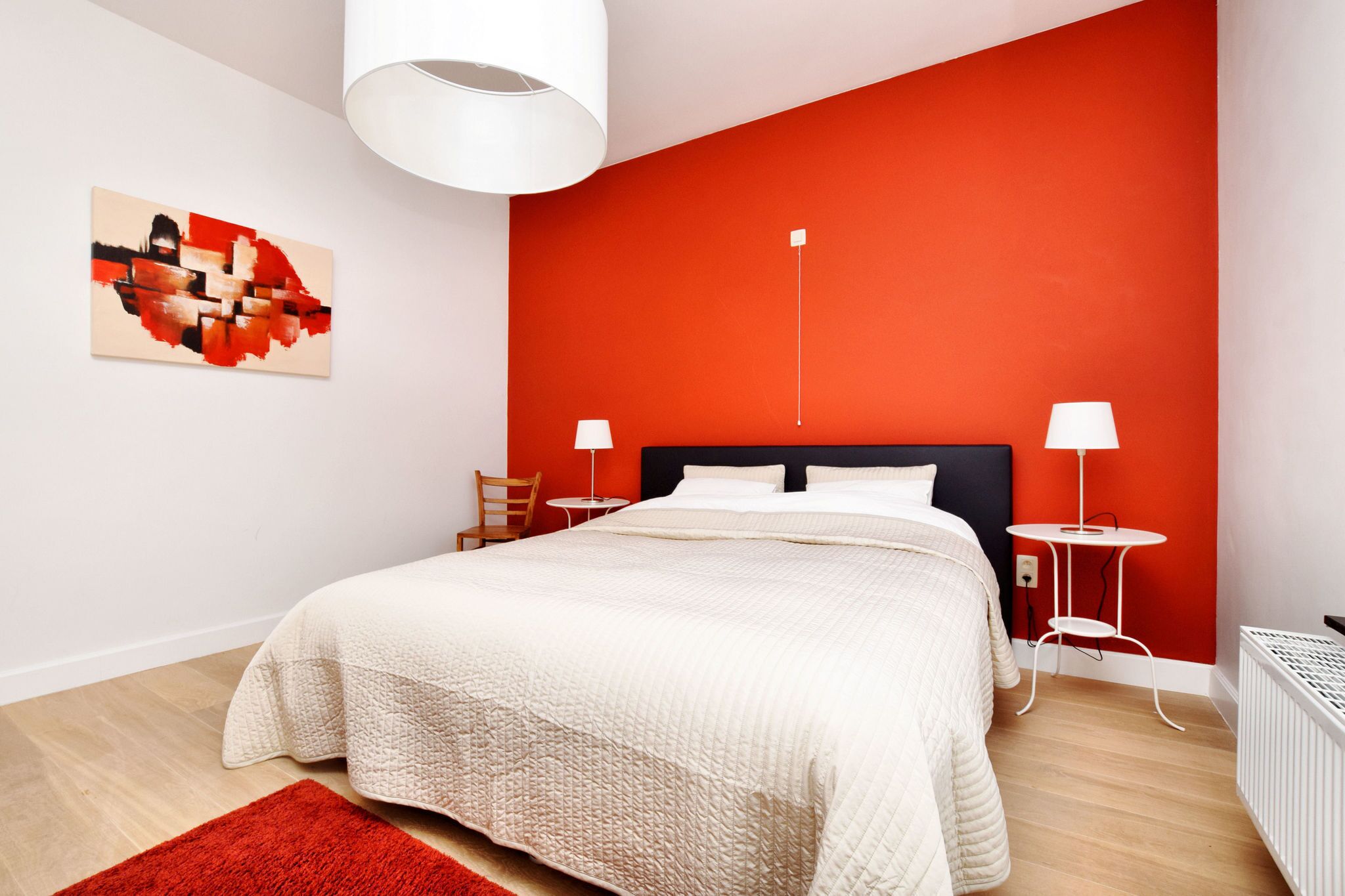 A modern bedroom with a large bed, a red accent wall, a white ceiling lamp, and a small side table with a lamp.