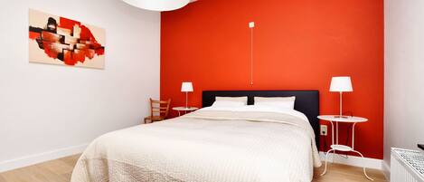 A modern bedroom with a large bed, a red accent wall, a white ceiling lamp, and a small side table with a lamp.