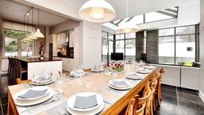 Un espace de salle à manger moderne, avec une longue table en bois dressée pour un repas, de grandes fenêtres et un mur d’accent en pierre.