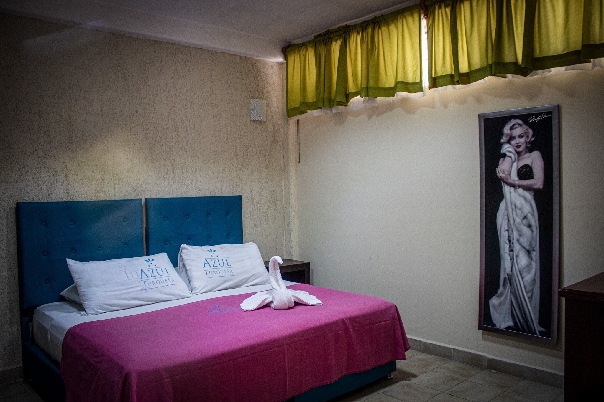 A hotel room with a bed, pillows, a decorative swan, a green curtain, and a framed portrait on the wall.