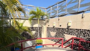 Piscina circular com design em mosaico, cercada por palmeiras e um guarda-corpo vermelho.