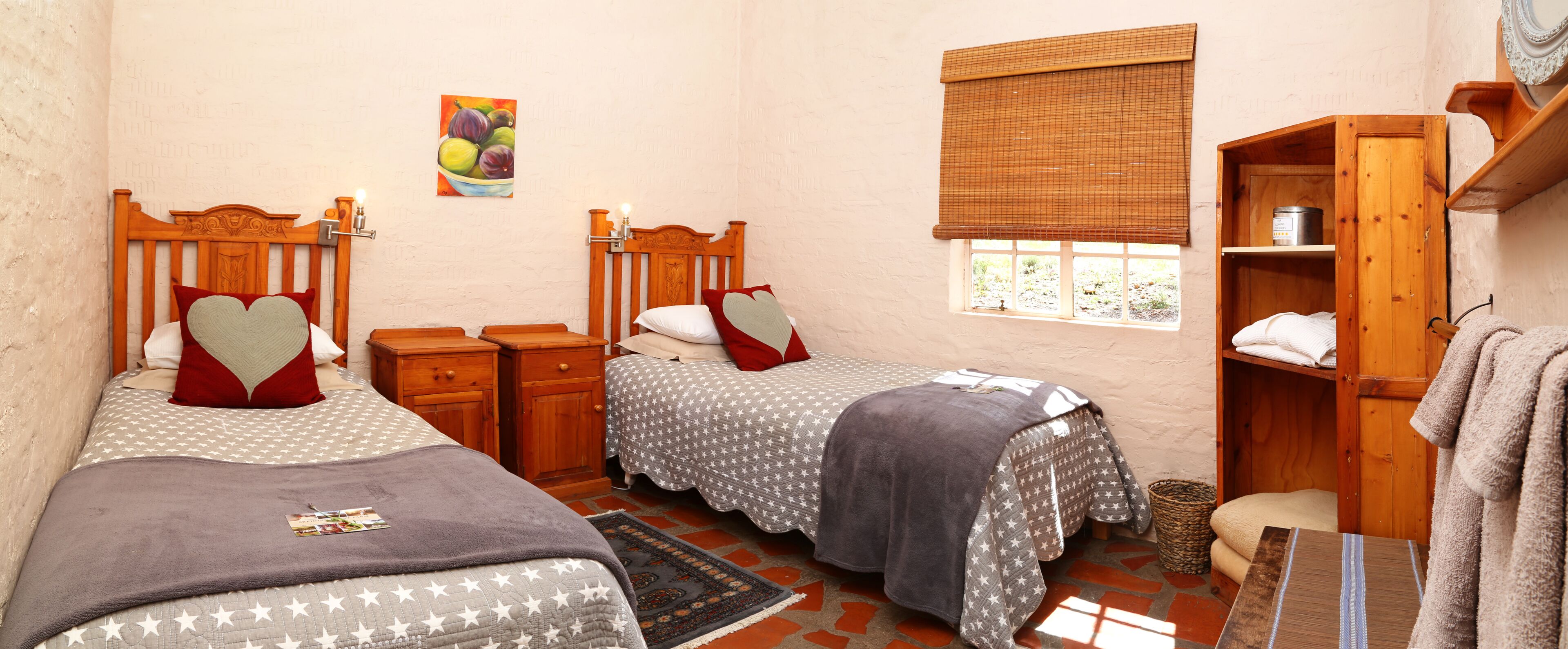 A bedroom with two beds, wooden headboards, a window with a blind, a bedside table with a lamp, a wall-mounted painting, and a wooden cabinet with towels.