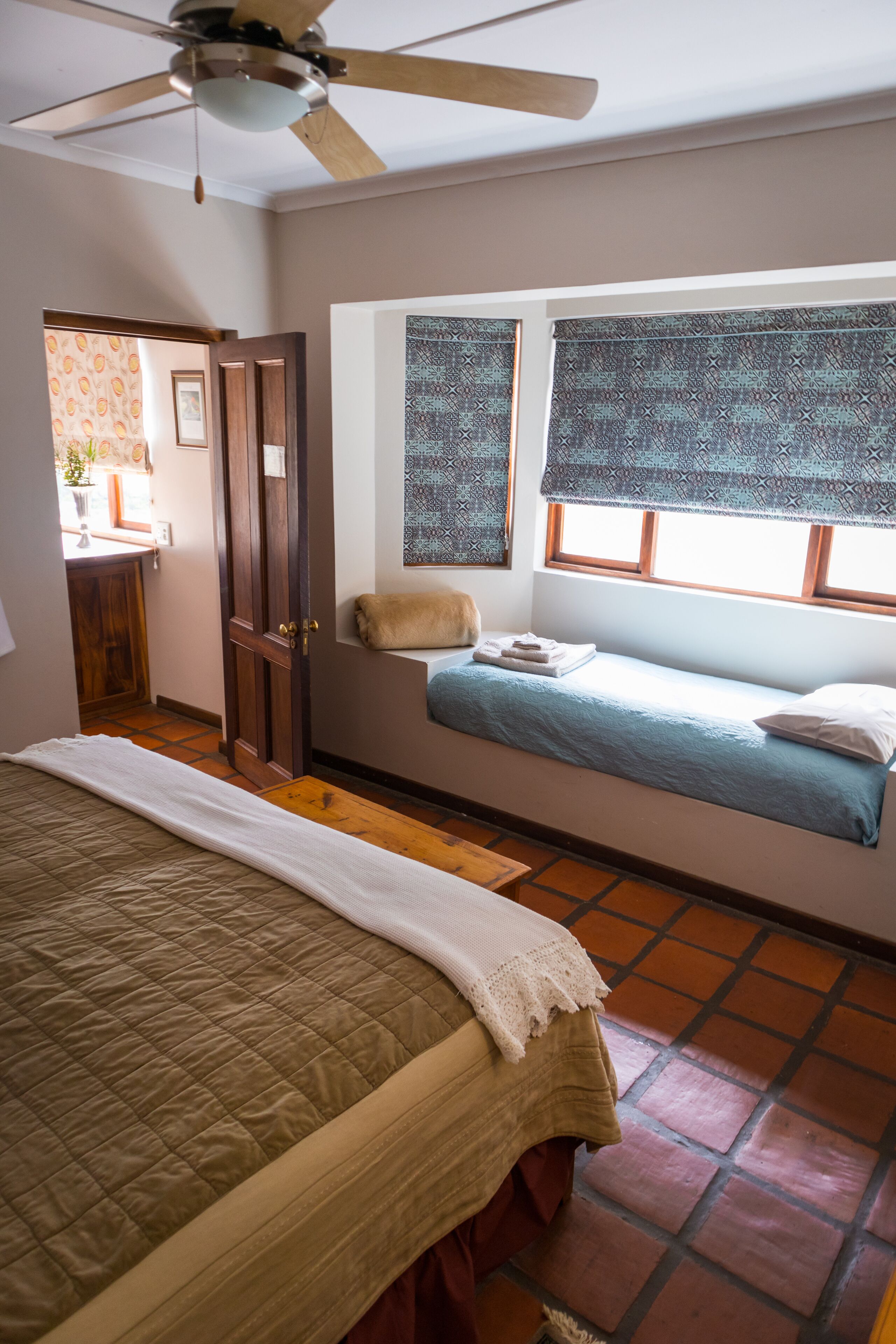 A bedroom with a bed, a window with patterned curtains, a ceiling fan, and a wooden door.