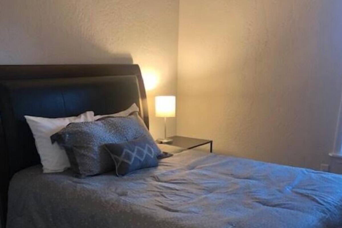 A neatly made bed with a dark headboard, a bedside table with a lamp, and a plain wall.