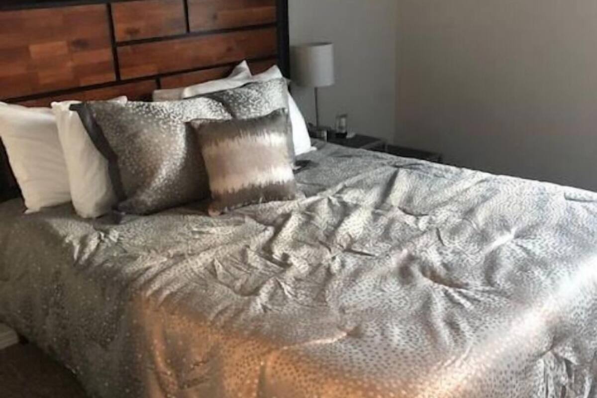 A neatly made bed with a patterned bedspread and several pillows, a wooden headboard, and a bedside table with a lamp.