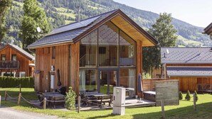 A wooden cabin with a large glass front, wooden deck, and a view of a mountainous landscape.