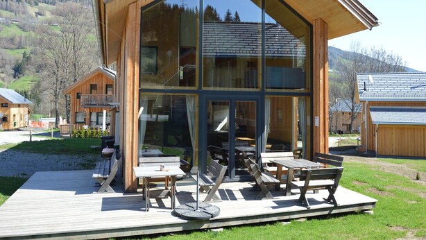 A modern wooden cabin with large glass windows, a deck with outdoor seating, and a view of the surrounding landscape.