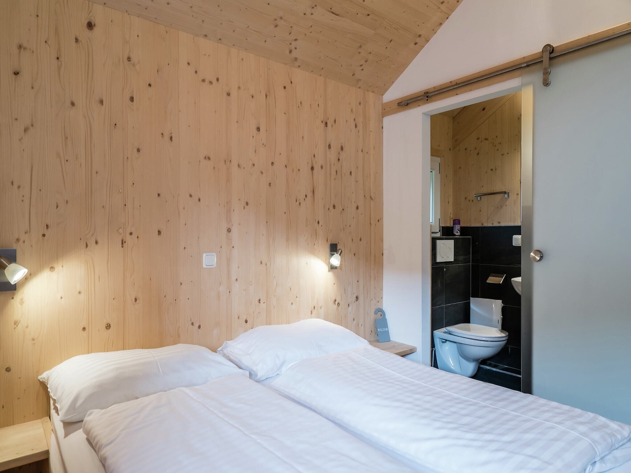 Una camera da letto con soffitto in legno, due letti con lenzuola bianche, un comodino, una lampada a parete e un bagno visibile attraverso una porta aperta.