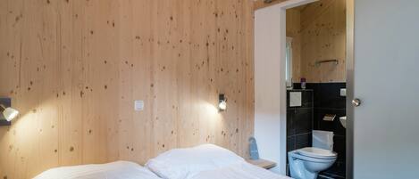 Una camera da letto con soffitto in legno, due letti con lenzuola bianche, un comodino, una lampada a parete e un bagno visibile attraverso una porta aperta.