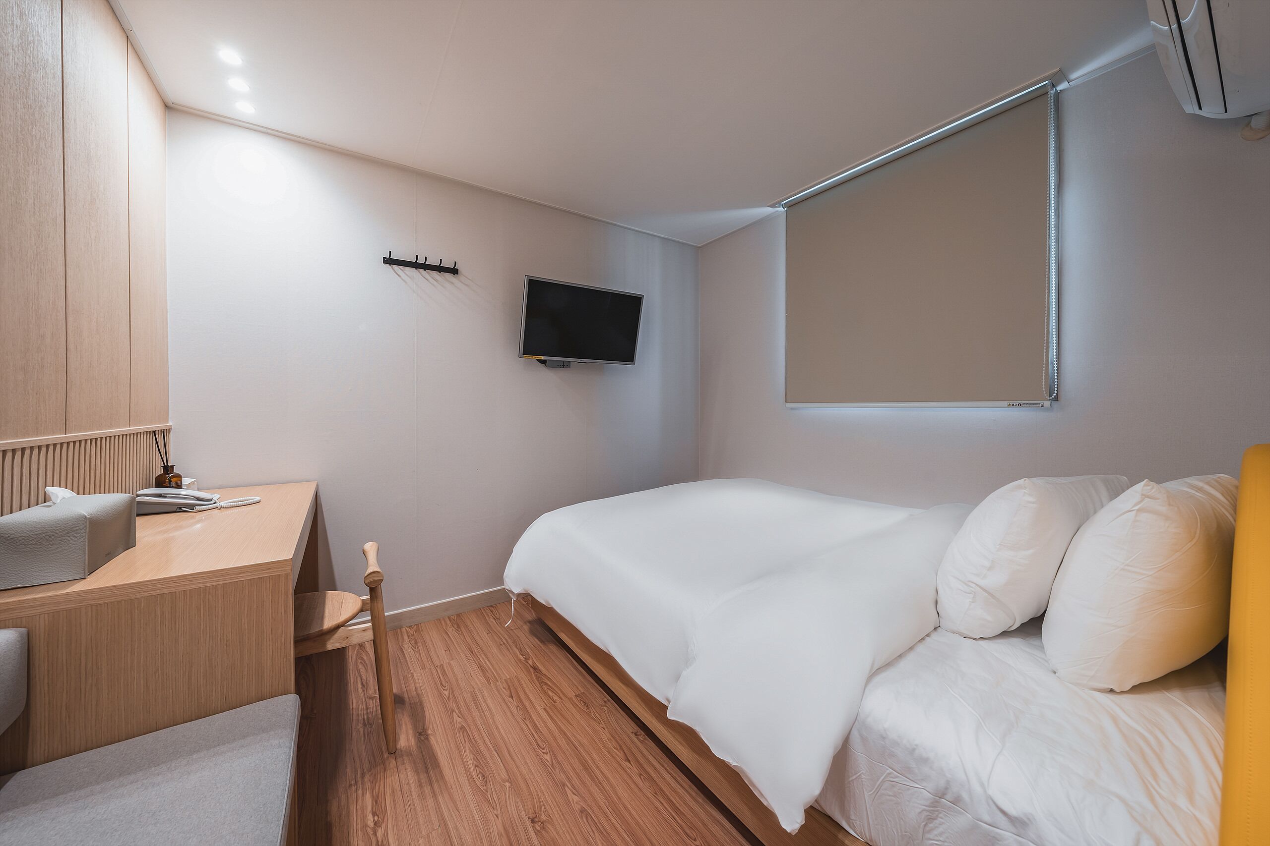 Een moderne slaapkamer met een houten bureau, een bed met witte beddengoed, een wandtelevisie en een raam met zonwering.