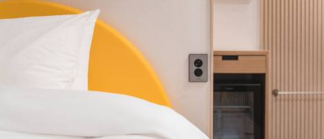 A bed with white bedding and a yellow headboard, a small wooden cabinet with a built-in oven, and a wall-mounted light switch.