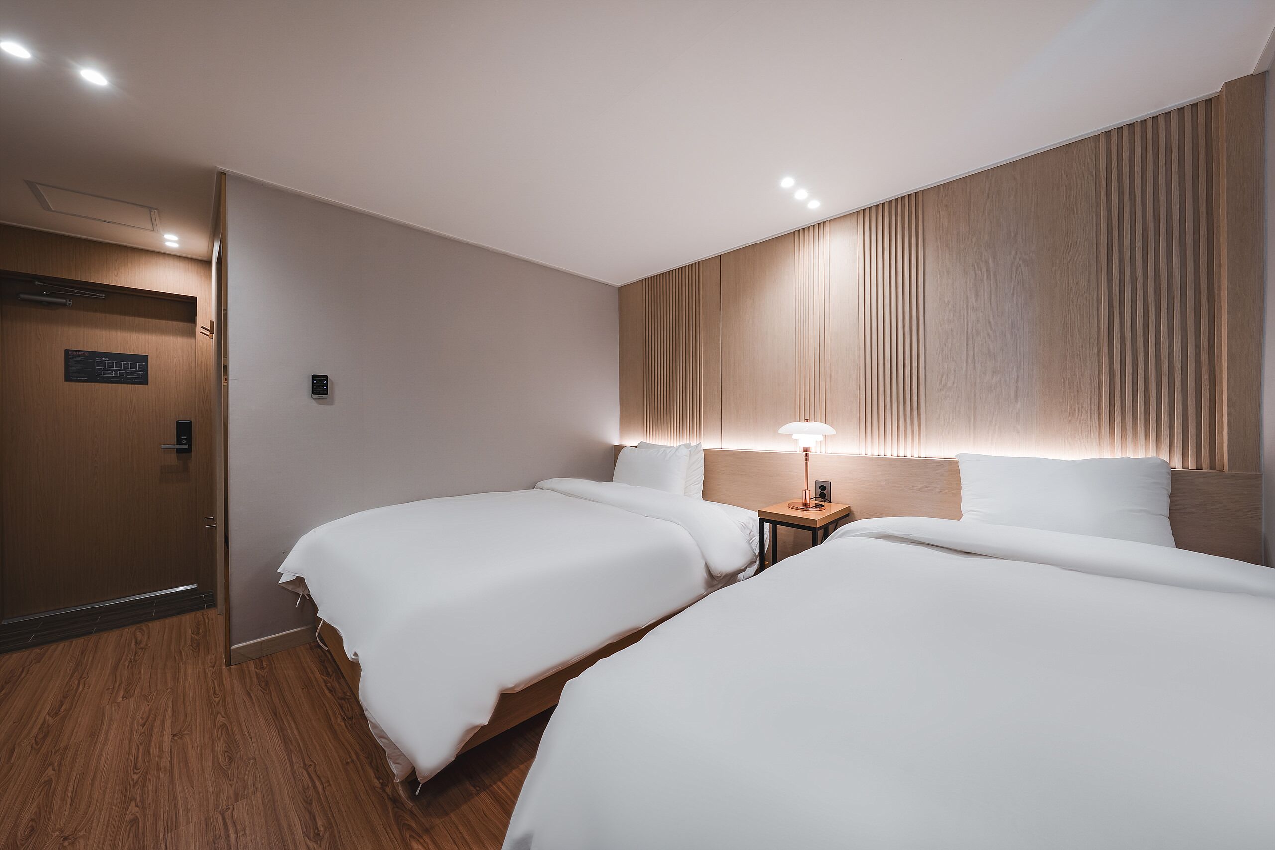 A hotel room with two beds, a wooden panel wall, a nightstand with a lamp, and a door with a control panel.