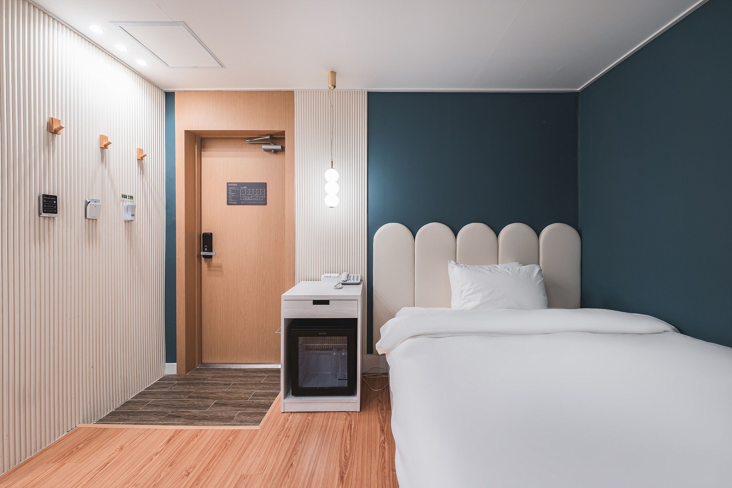 A hotel room with a bed, a bedside table with a phone, a heater, and a wall-mounted light fixture.