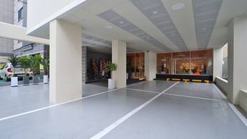 Una entrada de hotel moderna con zona cubierta, plantas en macetas y puerta de vidrio que da al interior.