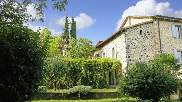 A stone building with a garden in front, featuring a hedge, a small tree, and a stone wall.