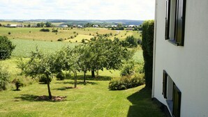 A view of a white building with shutters, overlooking a green field with trees and a distant village.