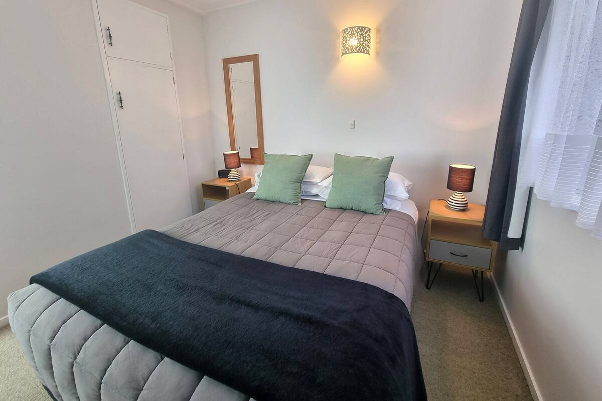 A bedroom with a bed, two bedside tables, a nightstand, a mirror, a wall-mounted lamp, and a window with curtains.