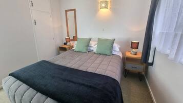 Une chambre à coucher comprenant un lit, deux tables de chevet, une commode, un miroir, une lampe fixée au mur et une fenêtre avec des rideaux.