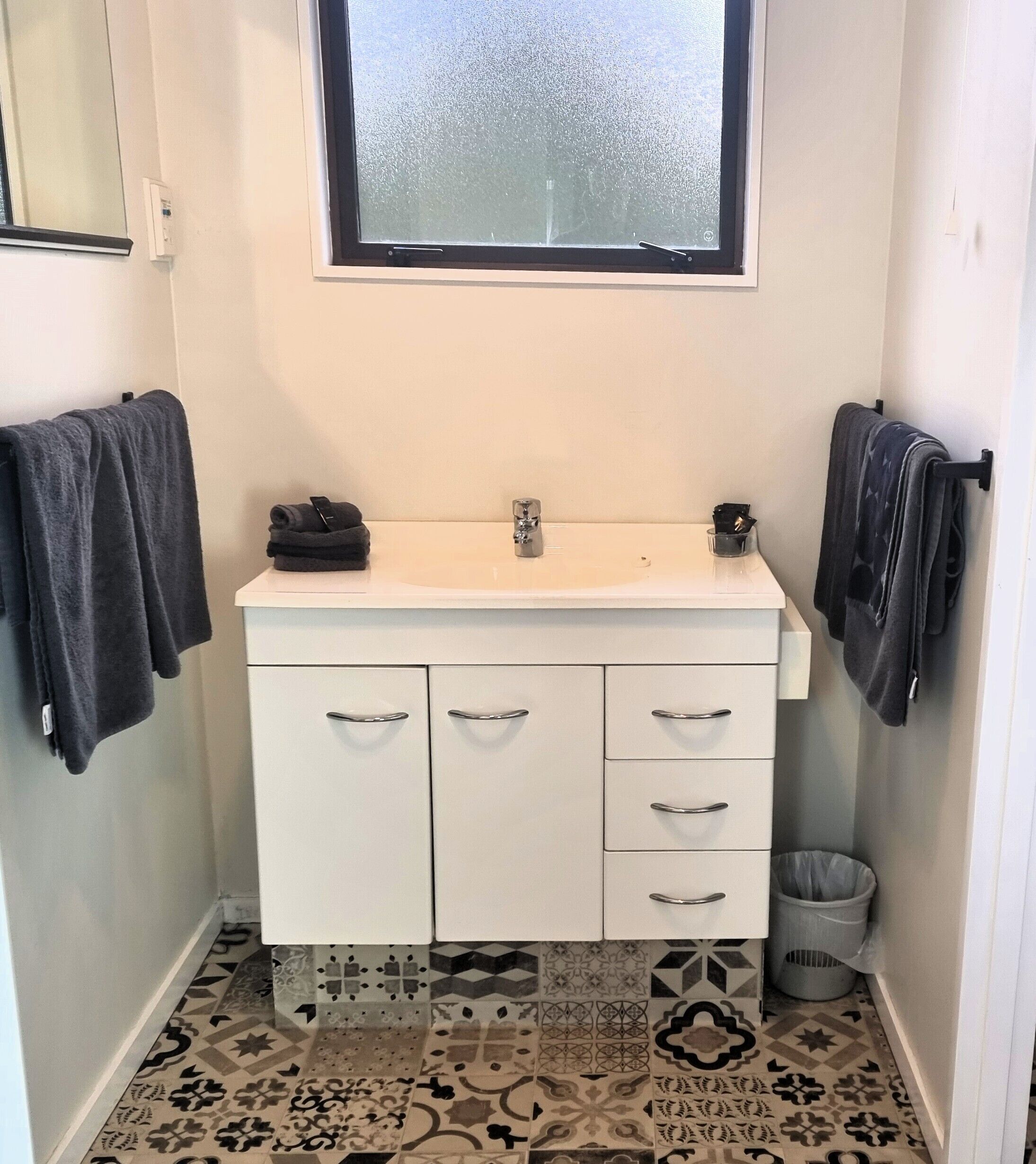 A small bathroom with a white sink, two drawers, a trash bin, and a window with frosted glass.