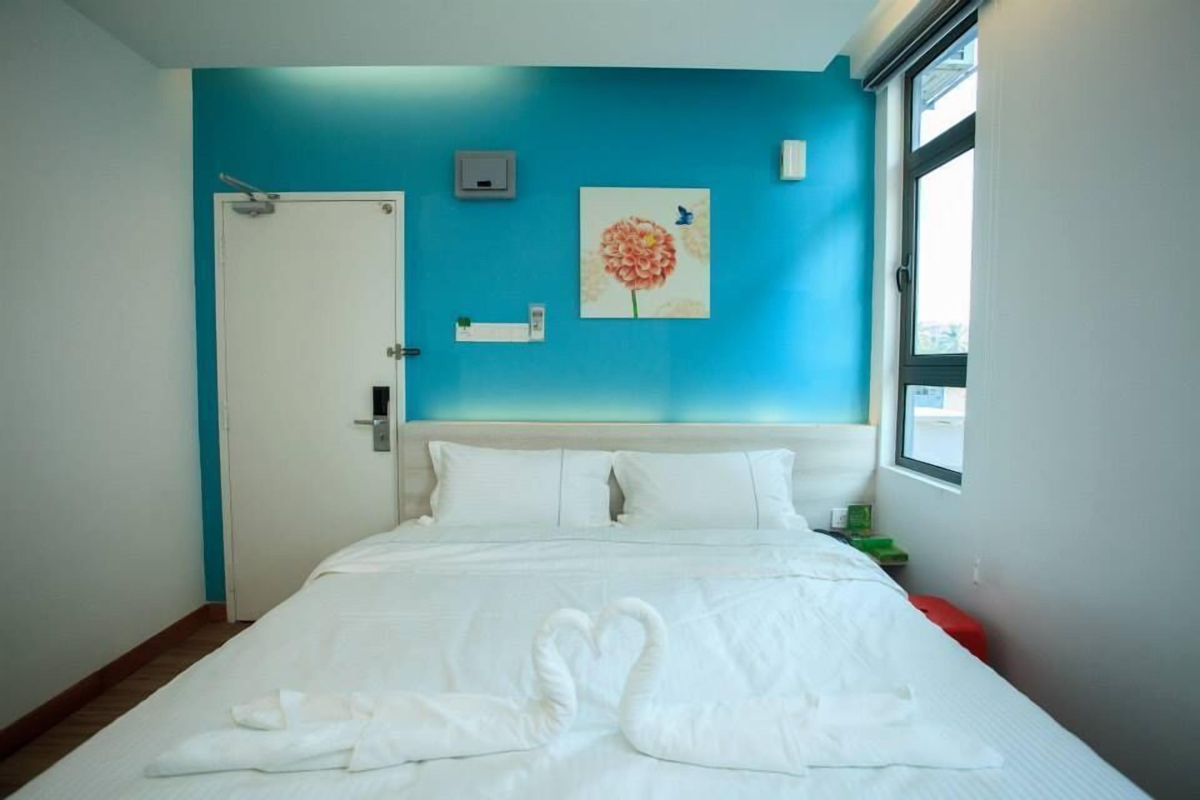 A hotel room with a bed, a door, a window, and a wall with a flower painting.