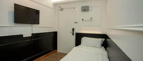 A small, modern hotel room with a single bed, a mounted TV, a door with a towel rack, and a small shelf with toiletries.